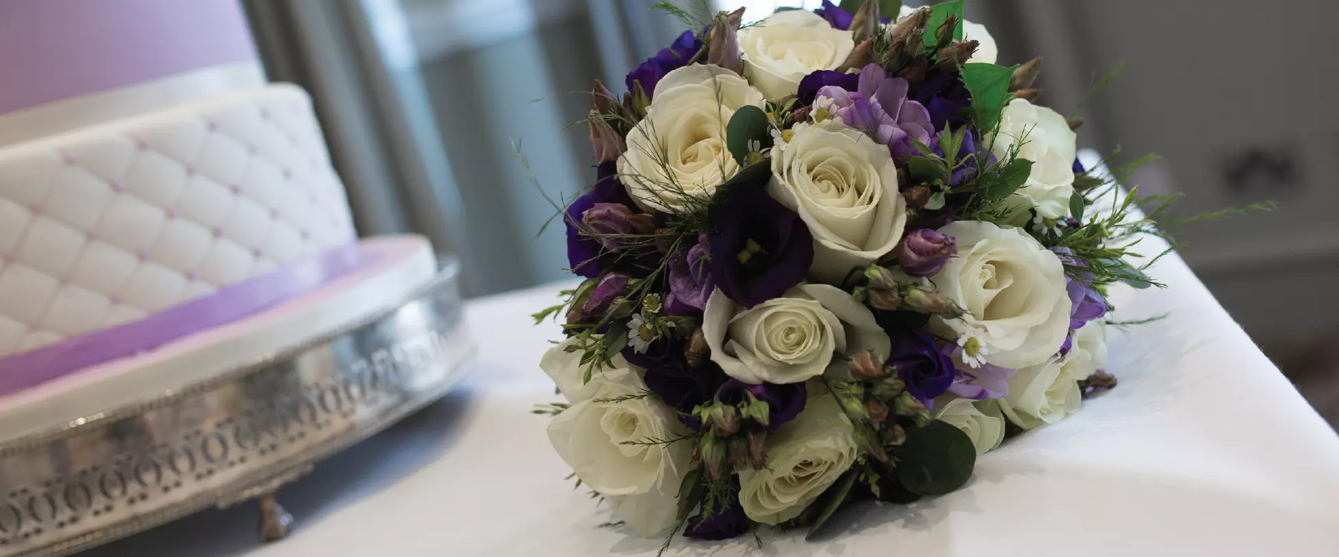 A bouquet of white and purple flowers on a white table sat next to a white and purple wedding cake..
